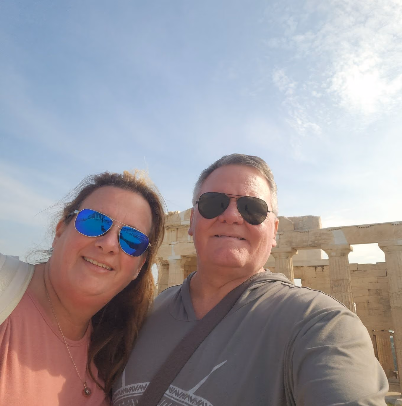 Smiling man and woman wearing sunglasses posing for selfie in front of ancient stone ruins under a blue sky.
