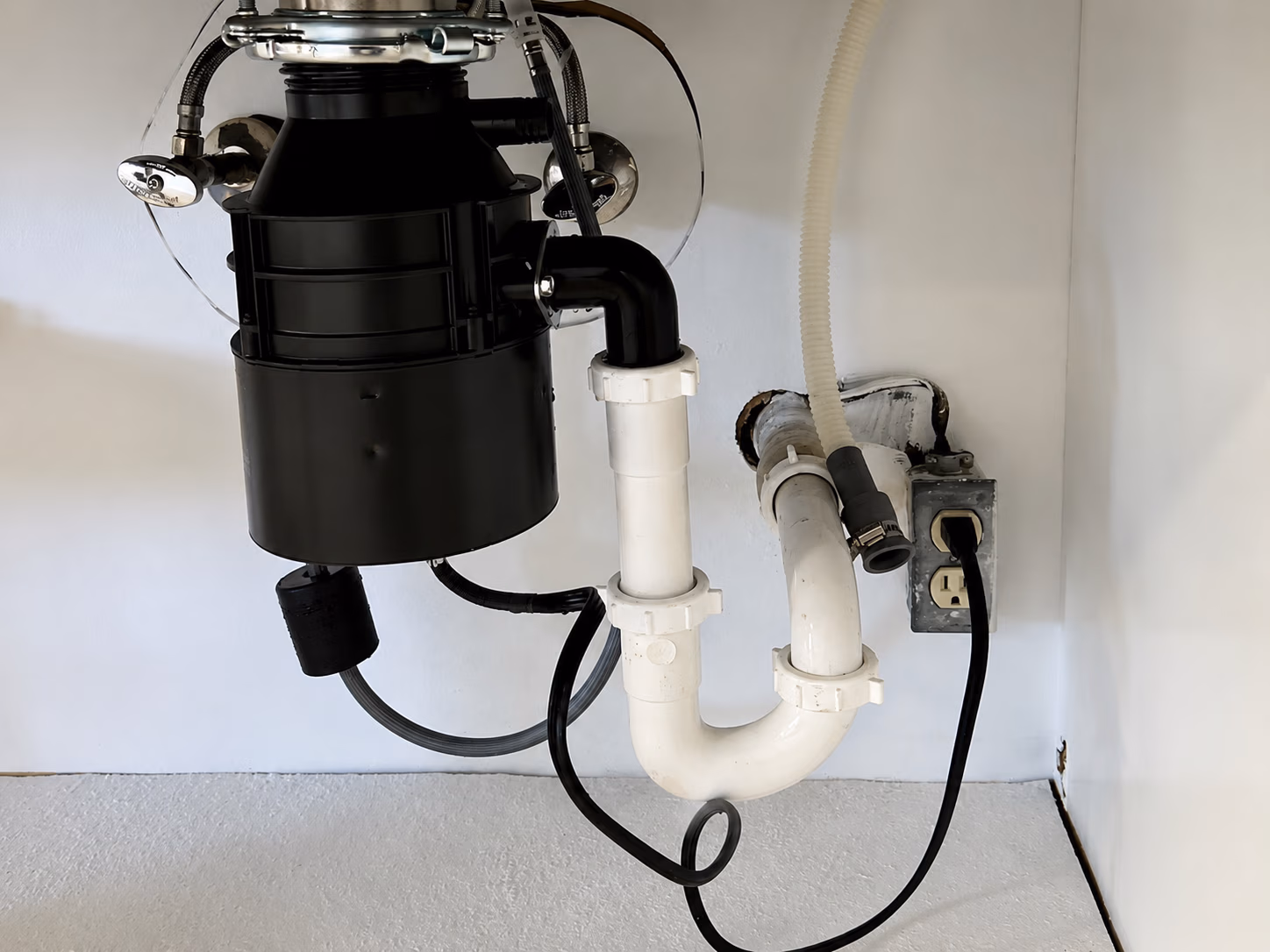 Under-sink plumbing with connected garbage disposal and drain pipes near an electrical outlet.
